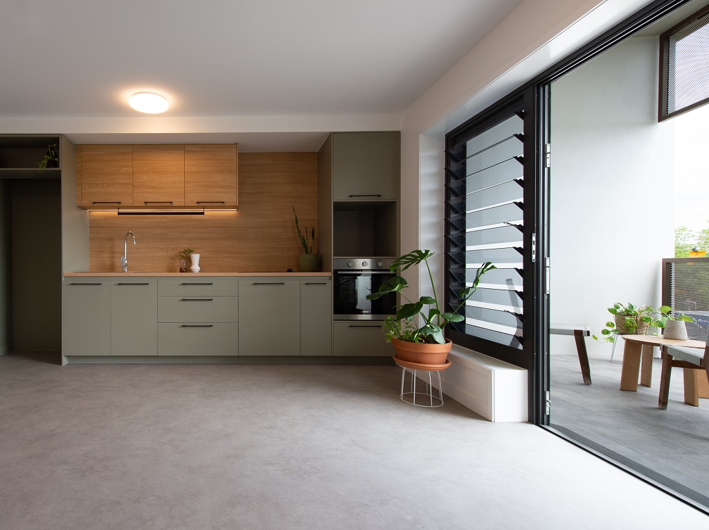 Interior view, showing the neutral-coloured kitchen with timber-look features, and large sliding doors and louvres looking out onto a generous balcony.