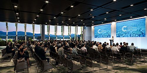 A Cairns Convention Centre room set up as a for a lecture with a speaker standing in front of an audience. Floor to ceiling windows overlooking the inlet and mountains. 
