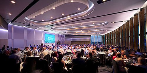 The Cairns Convention Centre Trinity room with tables of people at an event. The image shoes a presentation screen, over shaped ceiling lights, and floor to ceiling windows.