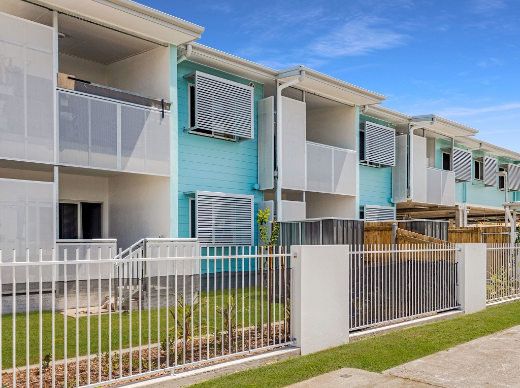 View from the footpath, showing private fenced yards and upper-level balconies. The building includes portions of weatherboards, painted bright aqua.