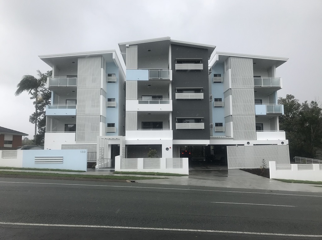 View from the street of a 4-storey development with secure carparking underneath. The windows and balconies include white aluminium screening. The building is painted an off-white colour, with light blue accents. 